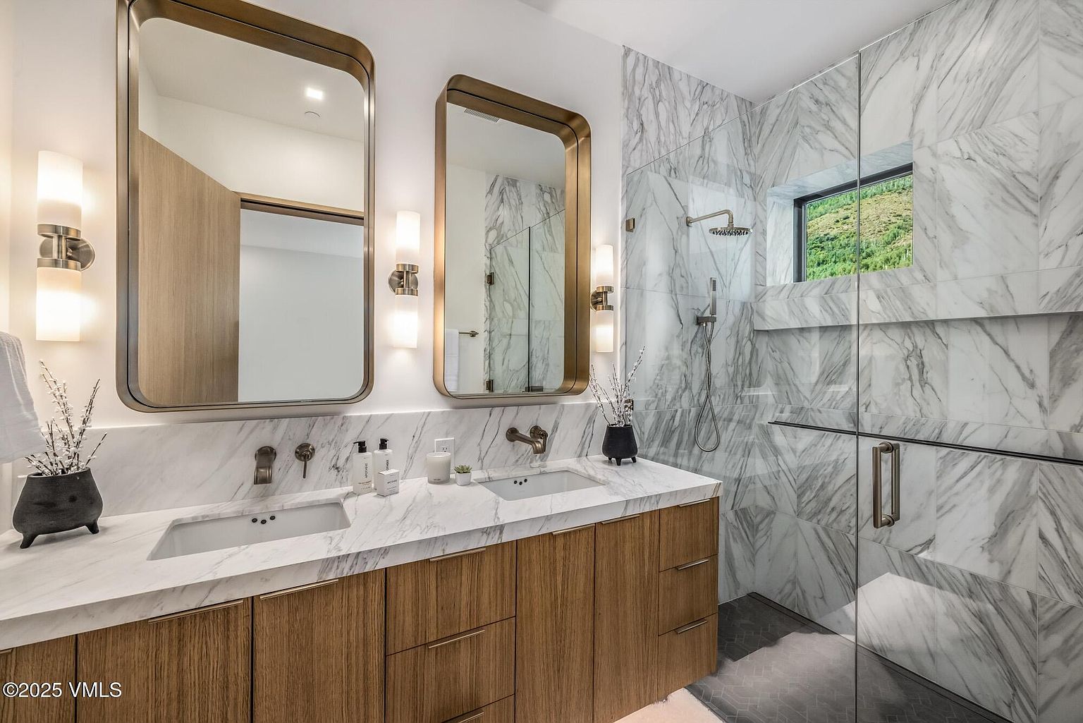 This modern primary bathroom features a dual-sink vanity with light wood cabinetry and a stone countertop. Above the vanity, two rectangular mirrors with metallic frames are flanked by wall-mounted light fixtures. To the right, a glass-enclosed shower area is finished with patterned stone wall tiles and includes a view of the outdoors through a rectangular window.