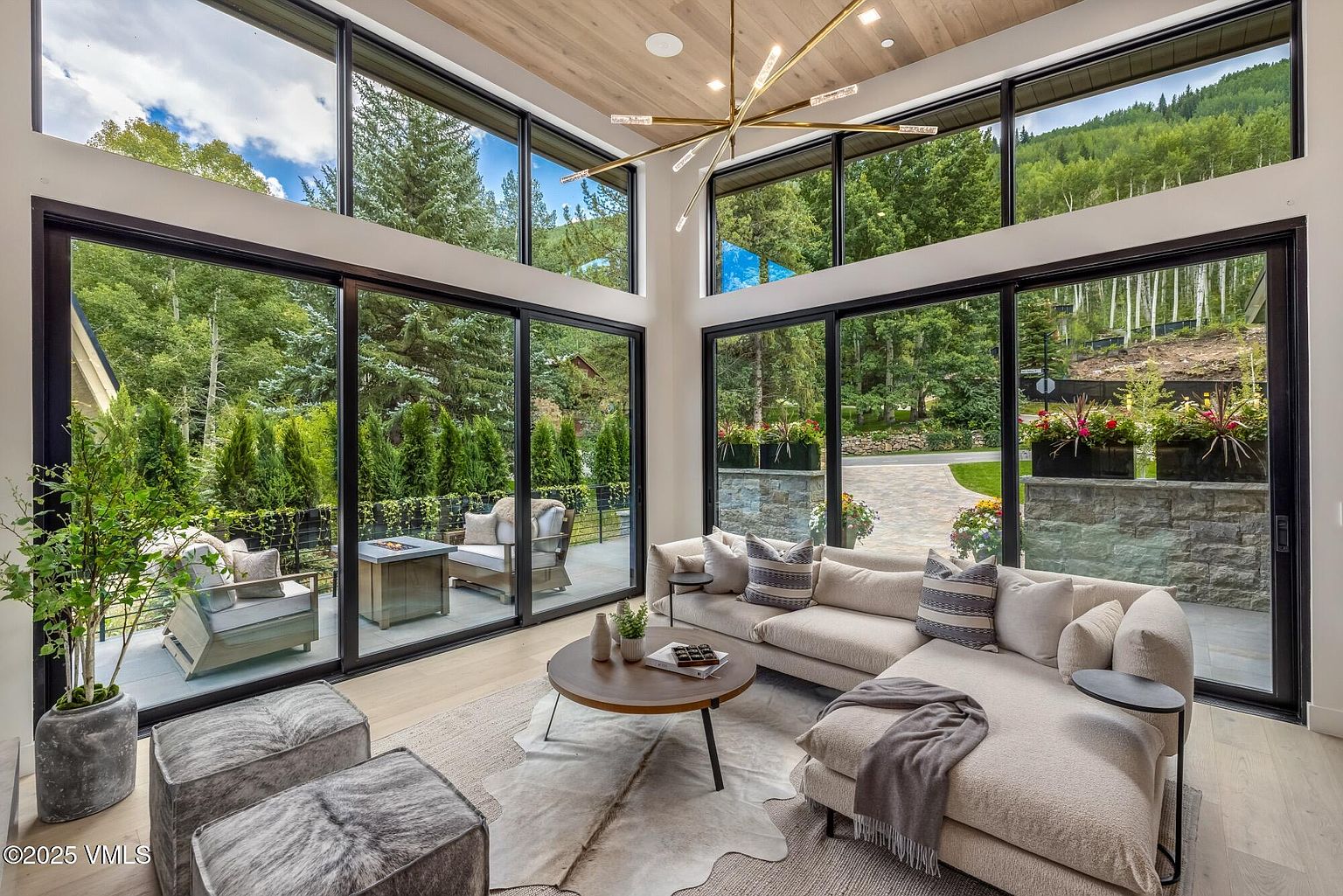 This bright and airy living room features expansive floor-to-ceiling windows that frame lush forest views and connect the space to an outdoor patio. The room is anchored by a large, light-toned sectional sofa, a circular wooden coffee table, and a textured area rug layered over a hide rug. A modern, multi-armed brass chandelier hangs from the light wood-paneled ceiling, creating a sophisticated and inviting atmosphere.