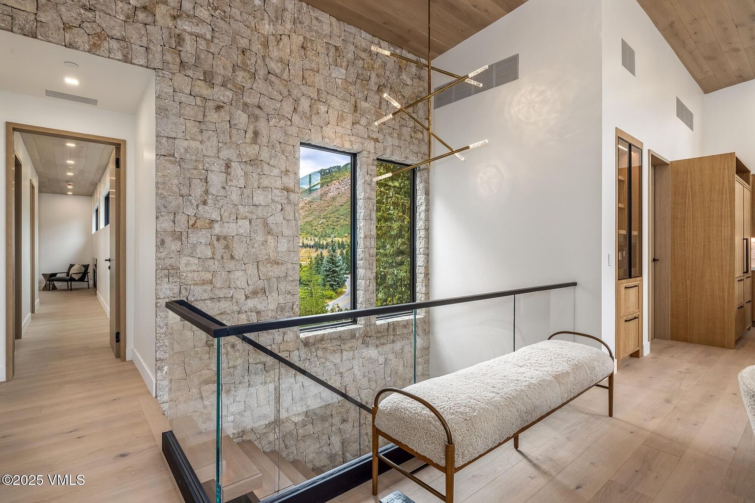 This elegant landing area features a striking stone-clad wall that frames two tall windows, offering a scenic view of the surrounding landscape. A modern, multi-armed brass chandelier hangs from the light wood-paneled ceiling, while a textured bench sits beside a glass-paneled staircase railing. The space is finished with light wood flooring, creating a bright, sophisticated atmosphere that seamlessly connects the different levels of the home.