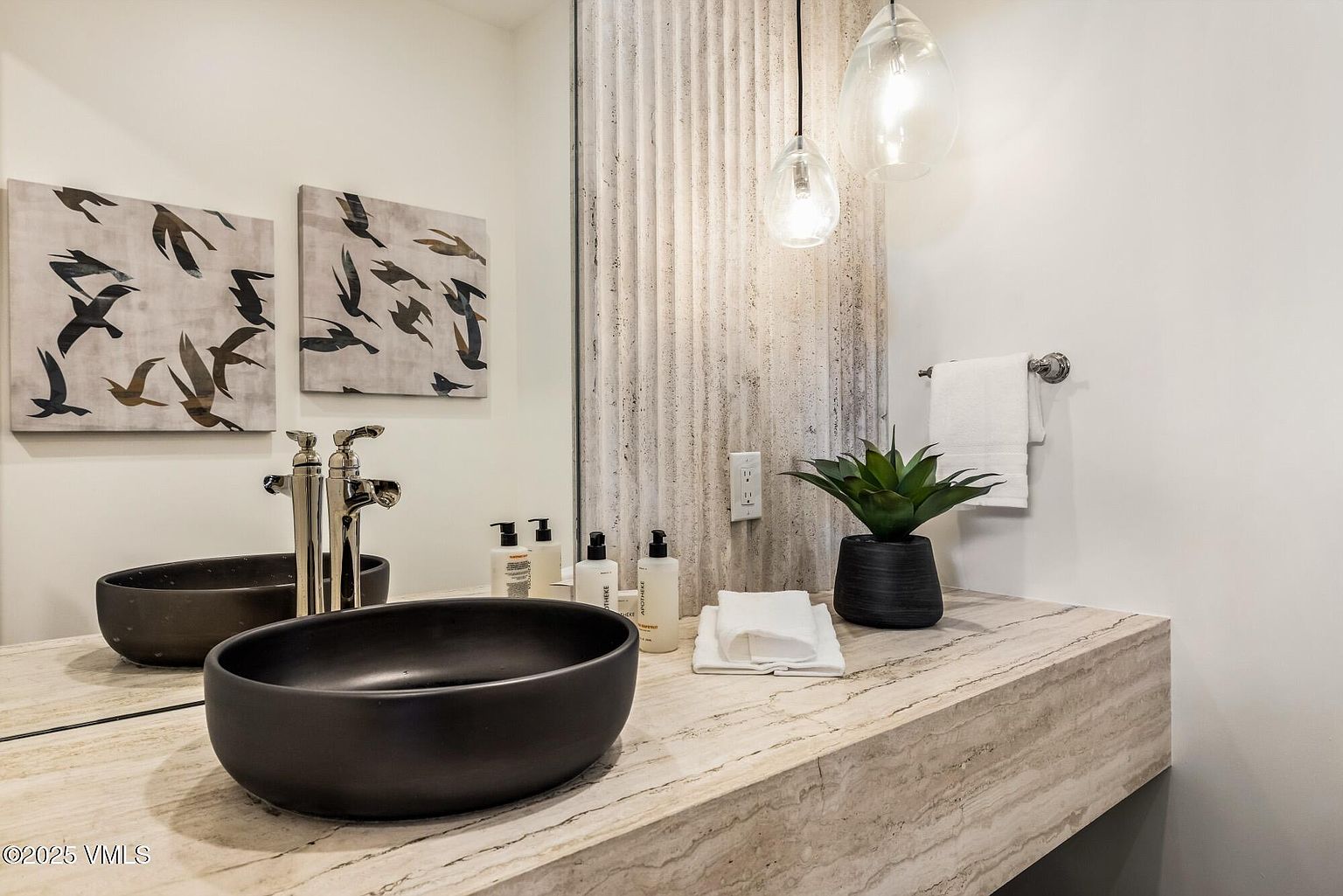 This modern powder room features a striking stone vanity with a prominent, textured stone backsplash that extends behind the mirror. A sleek, matte black vessel sink sits atop the vanity, complemented by contemporary wall-mounted faucets and two elegant glass pendant lights. The space is accented by minimalist bird-themed artwork and a small potted plant, creating a sophisticated and tranquil atmosphere.