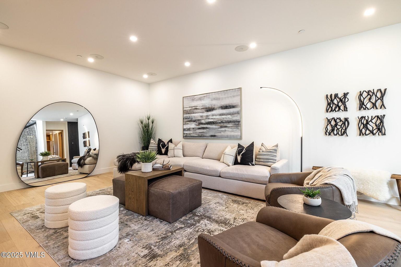 This contemporary living room features a neutral color palette with a large sectional sofa, two leather armchairs, and a pair of textured white ottomans. A prominent circular mirror reflects an adjacent room, while a large abstract painting hangs above the sofa, complemented by decorative wall art on the right. The space is anchored by a patterned area rug over light wood flooring, creating a sophisticated and inviting atmosphere.
