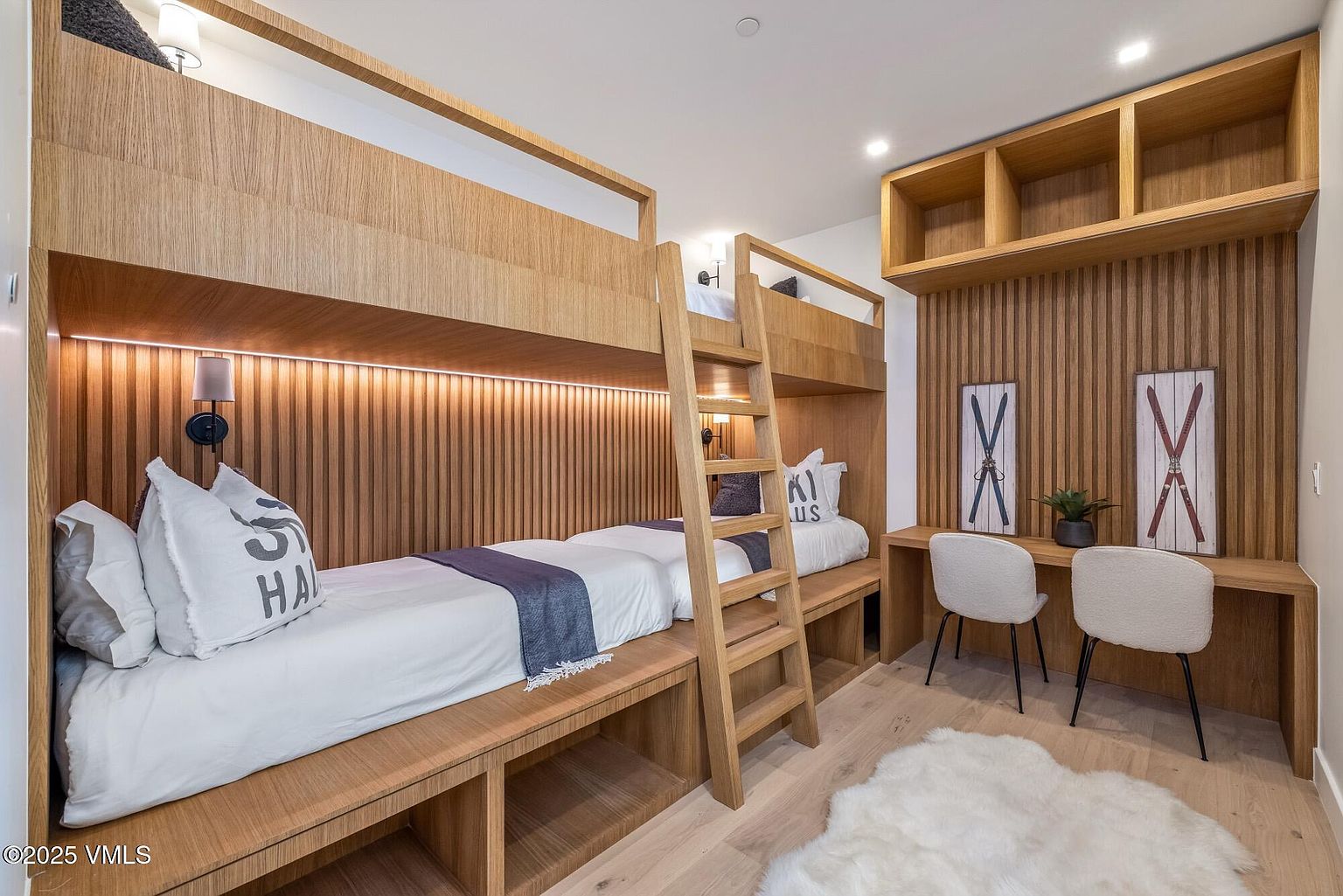 This cozy bedroom features custom-built bunk beds crafted from light wood, integrated with storage cubbies underneath. A matching desk area with two chairs is built into the wall, accented by vertical wood paneling and decorative ski-themed wall art. The room is finished with light wood flooring and a soft, white faux-fur rug, creating a warm and inviting mountain-inspired aesthetic.