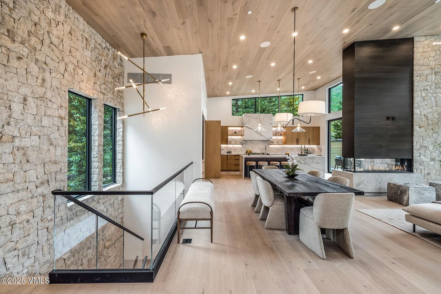 This open-concept living space features a dining area with a dark wood table and upholstered chairs, positioned adjacent to a kitchen with a stone-topped island. A prominent stone-clad chimney breast serves as a focal point, while a glass-railed staircase leads to a lower level. The room is characterized by light wood flooring, a matching wood-paneled ceiling, and large windows that provide a view of the surrounding trees.