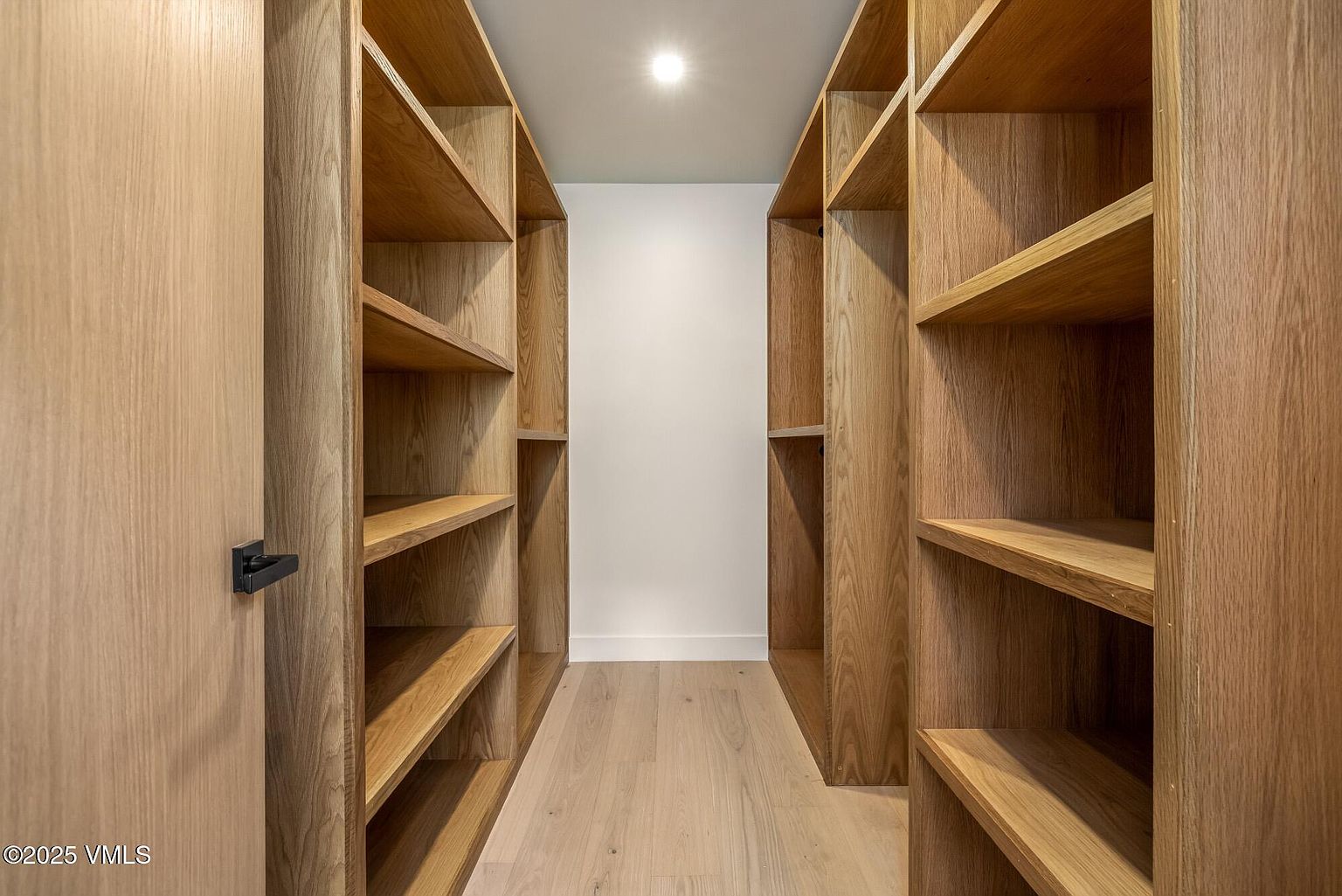 This walk-in closet features custom light wood shelving units lining both sides of a narrow, symmetrical corridor. The space is finished with light wood flooring and a clean, minimalist aesthetic, illuminated by a single recessed ceiling light. The perspective is centered, looking down the length of the closet toward a plain white back wall.