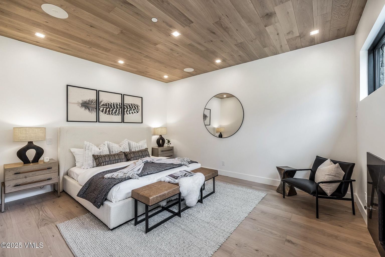 This primary bedroom features a sophisticated, modern aesthetic with light wood planking on the ceiling and matching light wood flooring. The room is anchored by a large, upholstered bed with layered bedding, flanked by two wooden nightstands with sculptural lamps. A circular mirror hangs on the white wall, while a black leather accent chair sits near a fireplace, creating a cozy and refined atmosphere.
