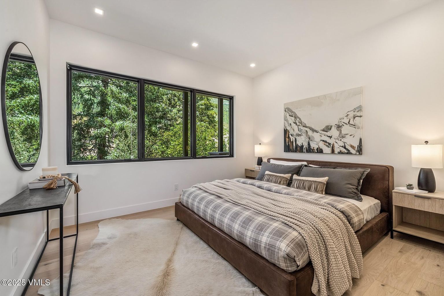 This modern bedroom features a low-profile bed with a brown upholstered frame, dressed in plaid bedding and a chunky knit throw. A large, multi-pane window provides a scenic view of the surrounding forest, while a round mirror and a slim console table add a contemporary touch to the left. The room is finished with light wood flooring, a textured area rug, and two bedside lamps that create a warm, inviting atmosphere.