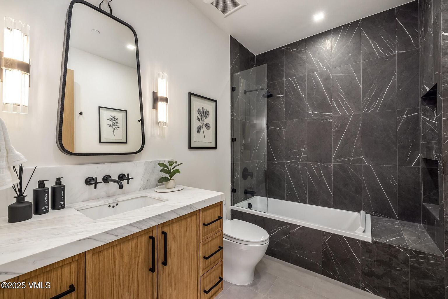 This modern bathroom features a vanity with a light wood finish and a stone countertop, paired with a sleek, dark-framed mirror and wall-mounted lighting. The shower area is enclosed by a glass panel and finished with dark, large-format stone tiles that extend into a built-in niche. The space is completed by a white toilet and neutral floor tiles, creating a clean, contemporary aesthetic.
