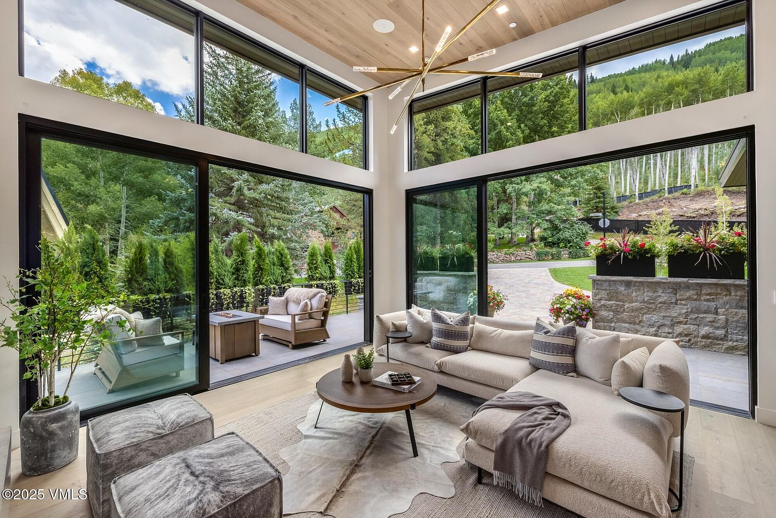 This bright and airy living room features expansive floor-to-ceiling windows that seamlessly blend the indoor space with the lush outdoor landscape. The room is anchored by a large, neutral-toned sectional sofa, a circular wooden coffee table, and two textured ottomans, all resting on a layered rug. A striking, modern gold-toned chandelier hangs from the light wood-paneled ceiling, adding a touch of elegance to the contemporary, nature-inspired design.