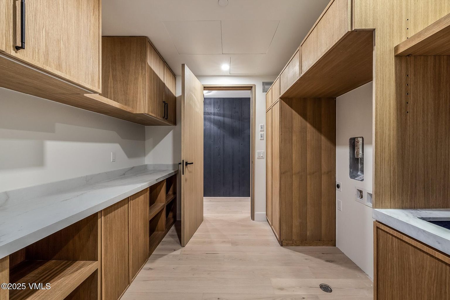 This functional laundry room features light wood cabinetry and a long stone countertop with an integrated sink. The space includes dedicated areas for laundry appliances, open shelving for storage, and light-toned wood flooring. A doorway leads into an adjacent room with dark-toned walls, creating a clean and modern aesthetic.