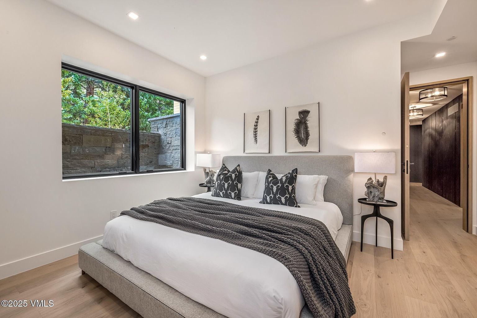 This contemporary bedroom features a neutral color palette with a plush, light-toned upholstered bed frame as the central focus. A large window provides natural light and a view of a stone retaining wall, while two bedside tables with unique sculptural lamps flank the bed. The room is accented by two framed feather-themed artworks above the headboard and light wood flooring, creating a serene and sophisticated atmosphere.