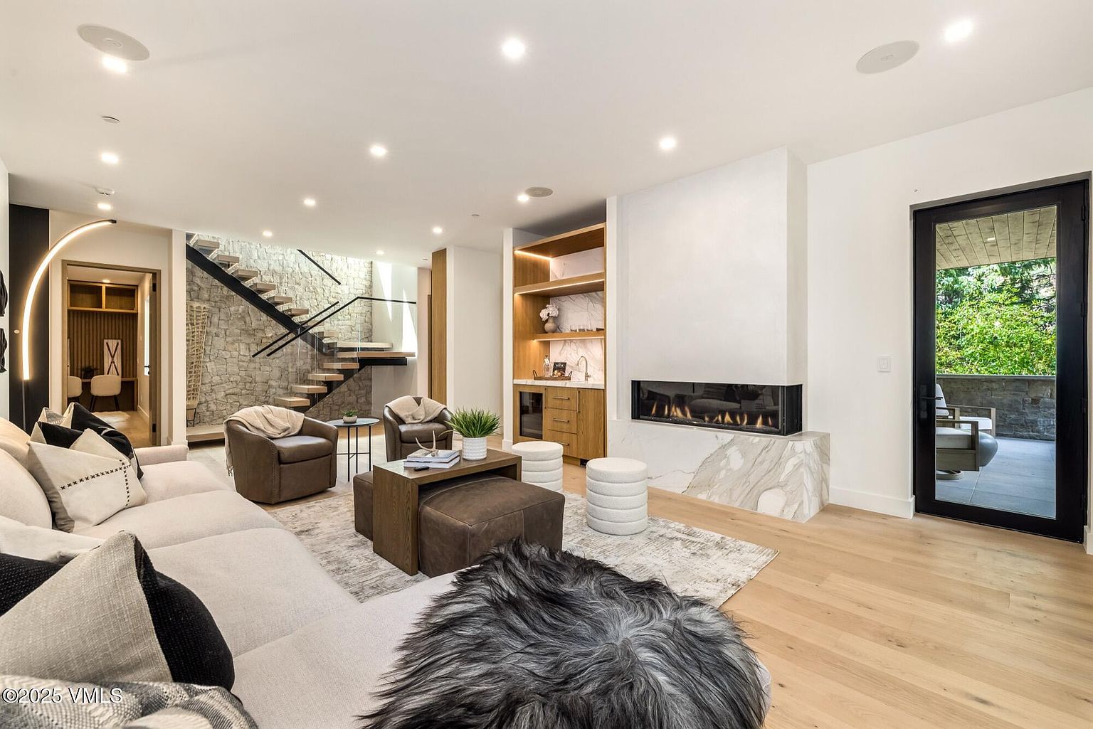 This contemporary living room features a light-toned sofa, a pair of leather armchairs, and a central coffee table set atop a textured area rug. A prominent feature is the linear fireplace set into a tall, light-veined stone surround, adjacent to a built-in wet bar with wood shelving. The space is anchored by a striking staircase with a stone-clad wall and glass railing, while a black-framed glass door provides a view to the outdoor patio.