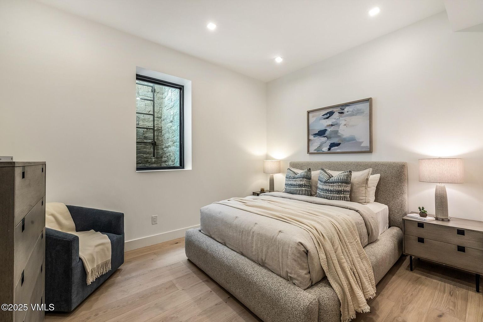This contemporary bedroom features a neutral color palette with a plush upholstered bed frame and light wood flooring. A dark blue armchair sits in the corner near a window that looks out onto a stone-lined light well, while a piece of abstract wall art hangs above the bed. The room is illuminated by recessed ceiling lights and two bedside lamps, creating a calm and inviting atmosphere.