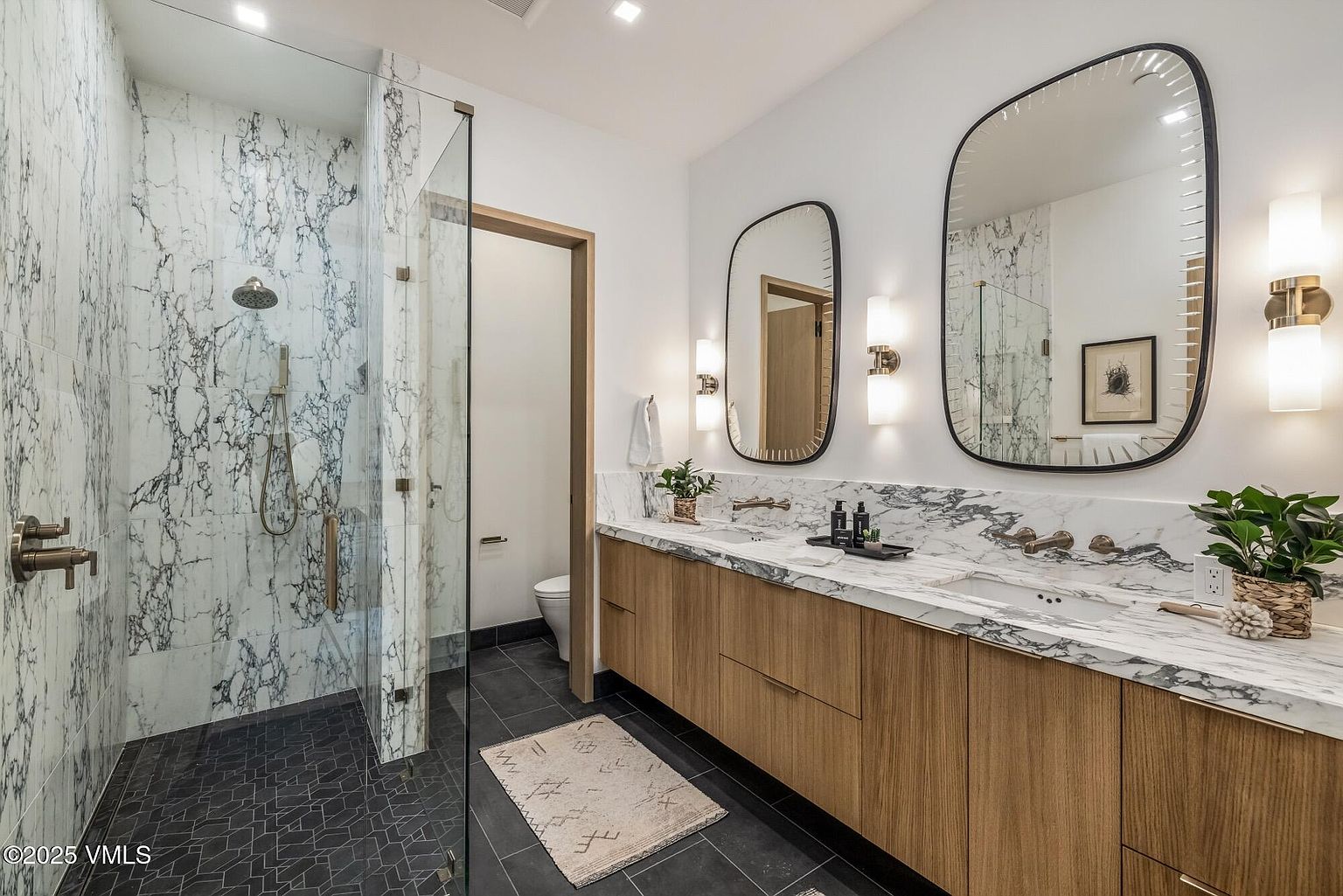 This modern primary bathroom features a double vanity with light wood cabinetry and a stone countertop, complemented by two distinct mirrors and wall-mounted light fixtures. To the left, a walk-in shower is enclosed by glass panels and finished with patterned stone walls and dark geometric floor tiles. The space is bright and contemporary, with a separate enclosed toilet area visible through a doorway.
