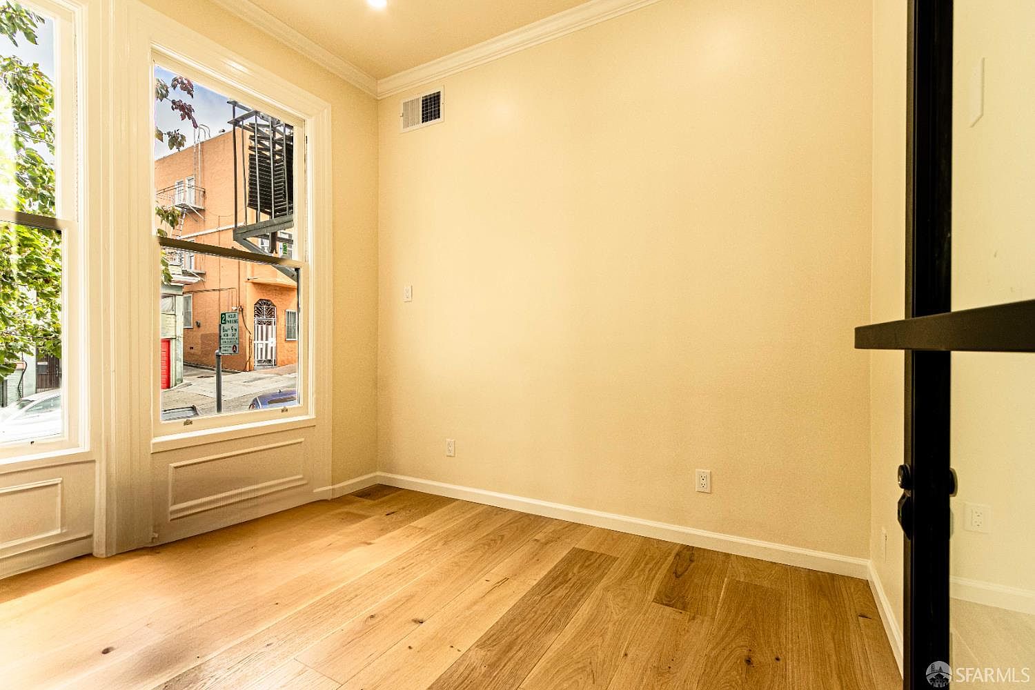 This bright, minimalist room features light wood flooring and neutral-toned walls, creating a clean and versatile space. A large window provides ample natural light and a view of the urban streetscape outside, including a fire escape. The room is framed by a dark-trimmed glass door on the right, adding a modern architectural contrast to the otherwise simple interior.