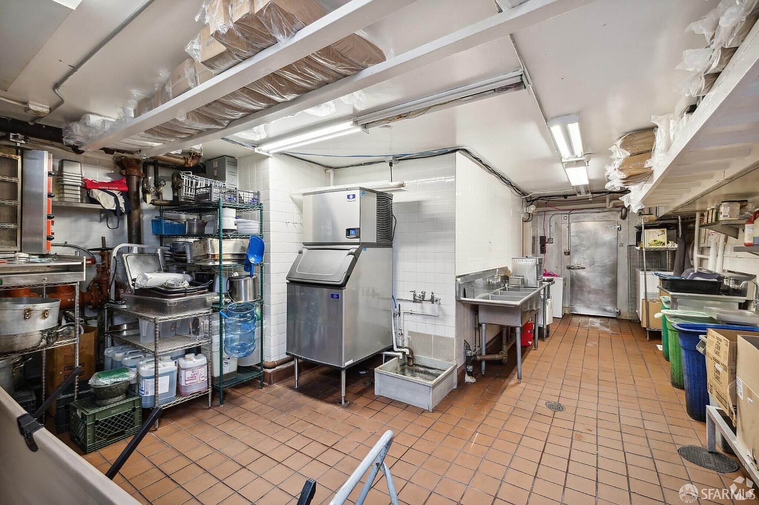 This image depicts a commercial-grade kitchen workspace characterized by industrial utility and functional storage. The room features tiled flooring, a large stainless steel ice machine, a multi-basin sink, and metal shelving units packed with various kitchen supplies and containers. The perspective is a wide-angle shot looking down the length of the room toward a metal door at the far end, emphasizing the utilitarian nature of the space.