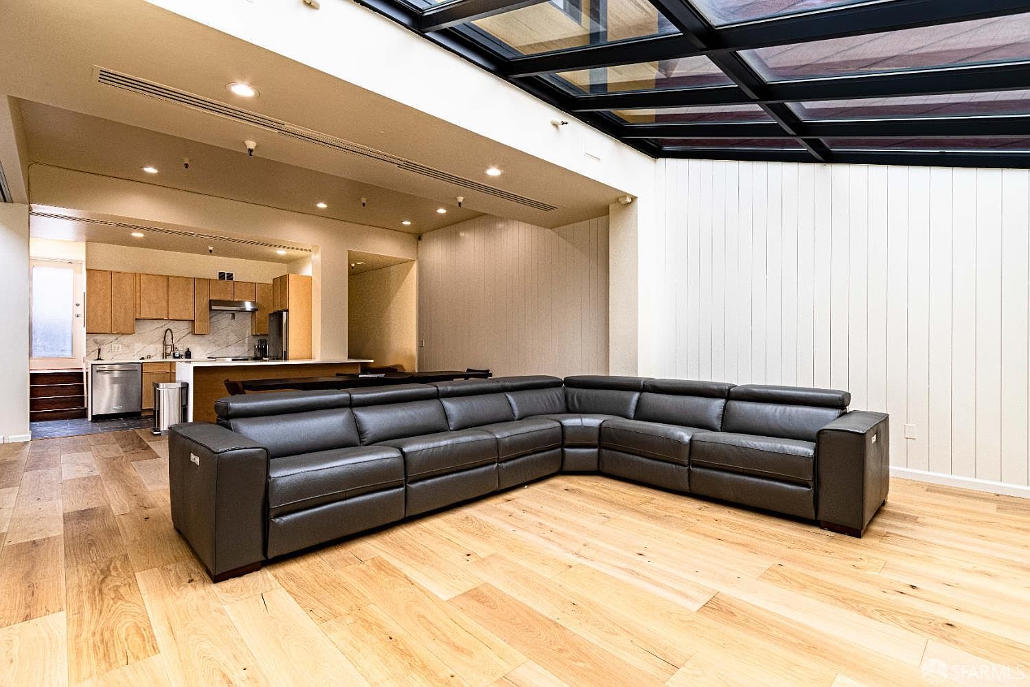 This open-concept living area features a large, dark leather sectional sofa set against a wall of vertical paneling. The space is defined by light-toned wood flooring and a dramatic glass skylight roof that floods the room with natural light. In the background, a kitchen with light wood cabinetry and a breakfast bar is visible, creating a seamless flow between the living and cooking spaces.