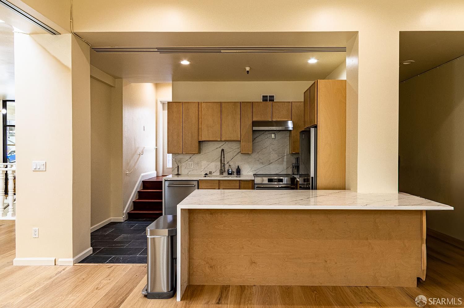 This kitchen features light wood cabinetry, a stone-topped island, and a matching stone backsplash. The space is equipped with stainless steel appliances and opens into a transition area with stairs leading to a lower level. The neutral color palette and clean lines create a bright, contemporary atmosphere.