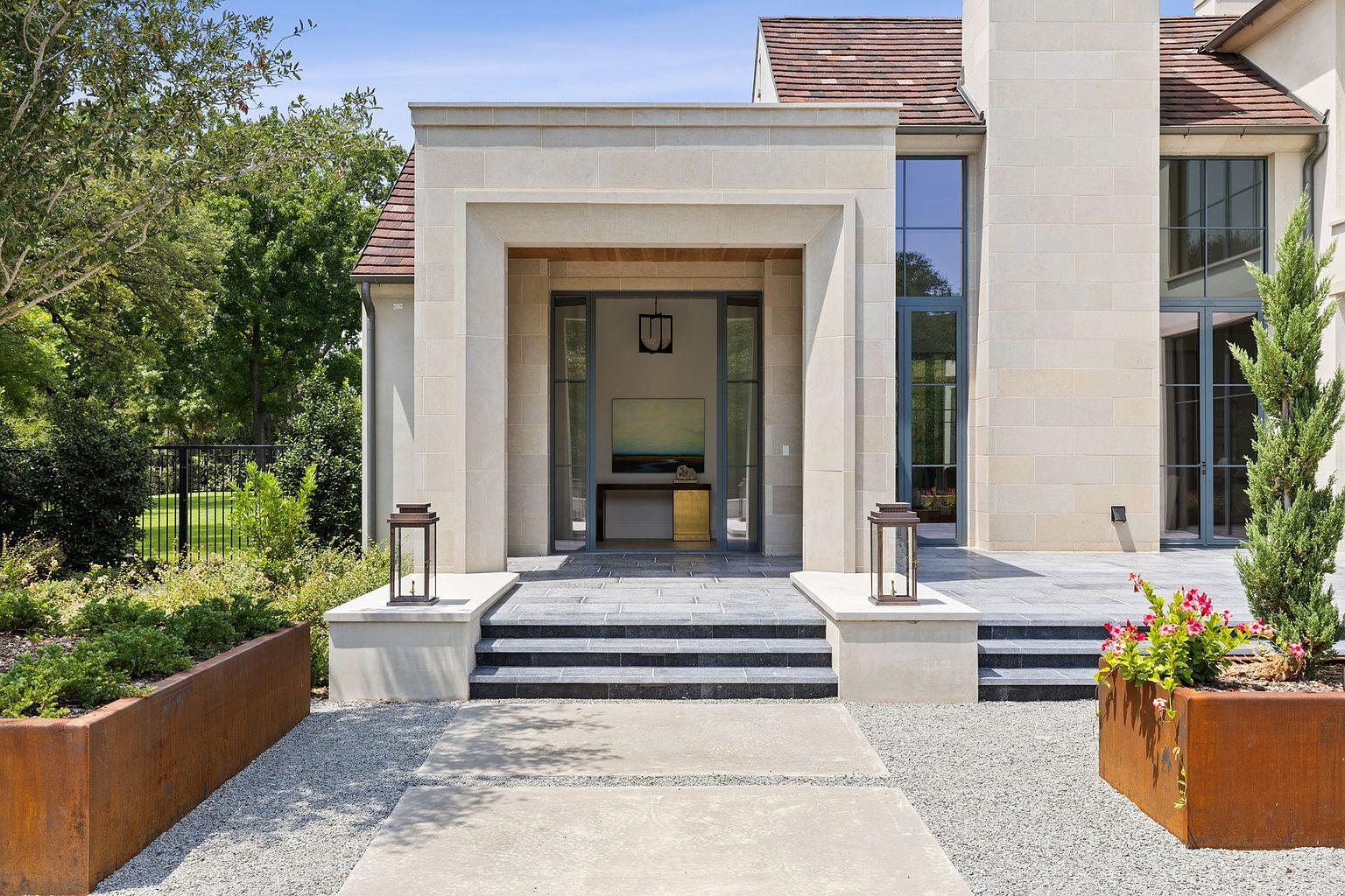 The image showcases a grand entryway to a modern home, featuring a stone facade, a covered porch with a dark-framed glass door, and a series of steps leading up to the entrance. Planters with greenery flank the walkway, adding a touch of nature to the architectural design. The overall impression is one of sophisticated elegance and curb appeal.