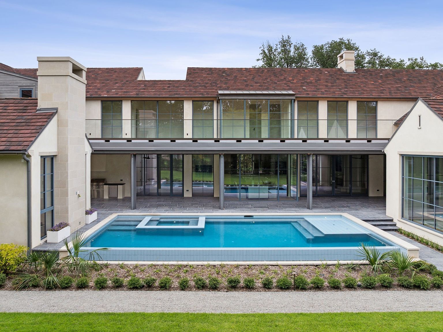 This image showcases a luxurious backyard featuring a modern pool and spa area. The pool has a unique design with integrated platforms and is surrounded by manicured landscaping and a gravel pathway. The house, with its large windows and covered patio, provides a stunning backdrop, creating an inviting and upscale outdoor living space.