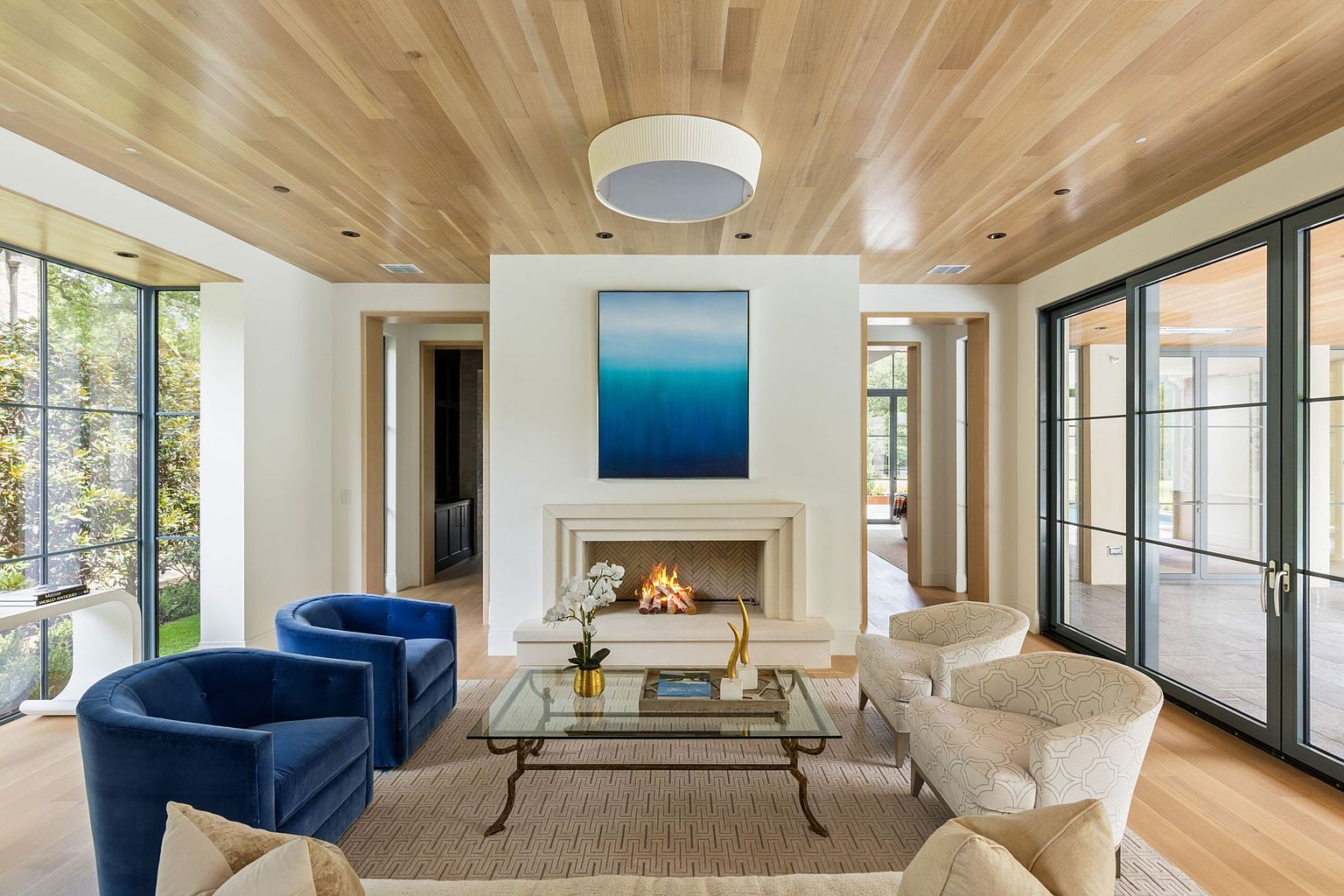This is a bright and airy living room featuring a fireplace with a blue painting above it, two blue armchairs, and two patterned armchairs. The room has large windows that let in plenty of natural light, and the ceiling is made of wood. The overall style is modern and sophisticated, creating a welcoming and comfortable atmosphere.