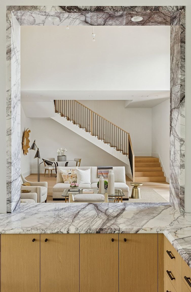 This interior shot showcases a bright and airy living room, viewed through a marble-framed opening. The space features a neutral-toned sofa, glass coffee table, and a staircase leading to an upper level. The design emphasizes clean lines and natural light, creating a welcoming and sophisticated atmosphere.