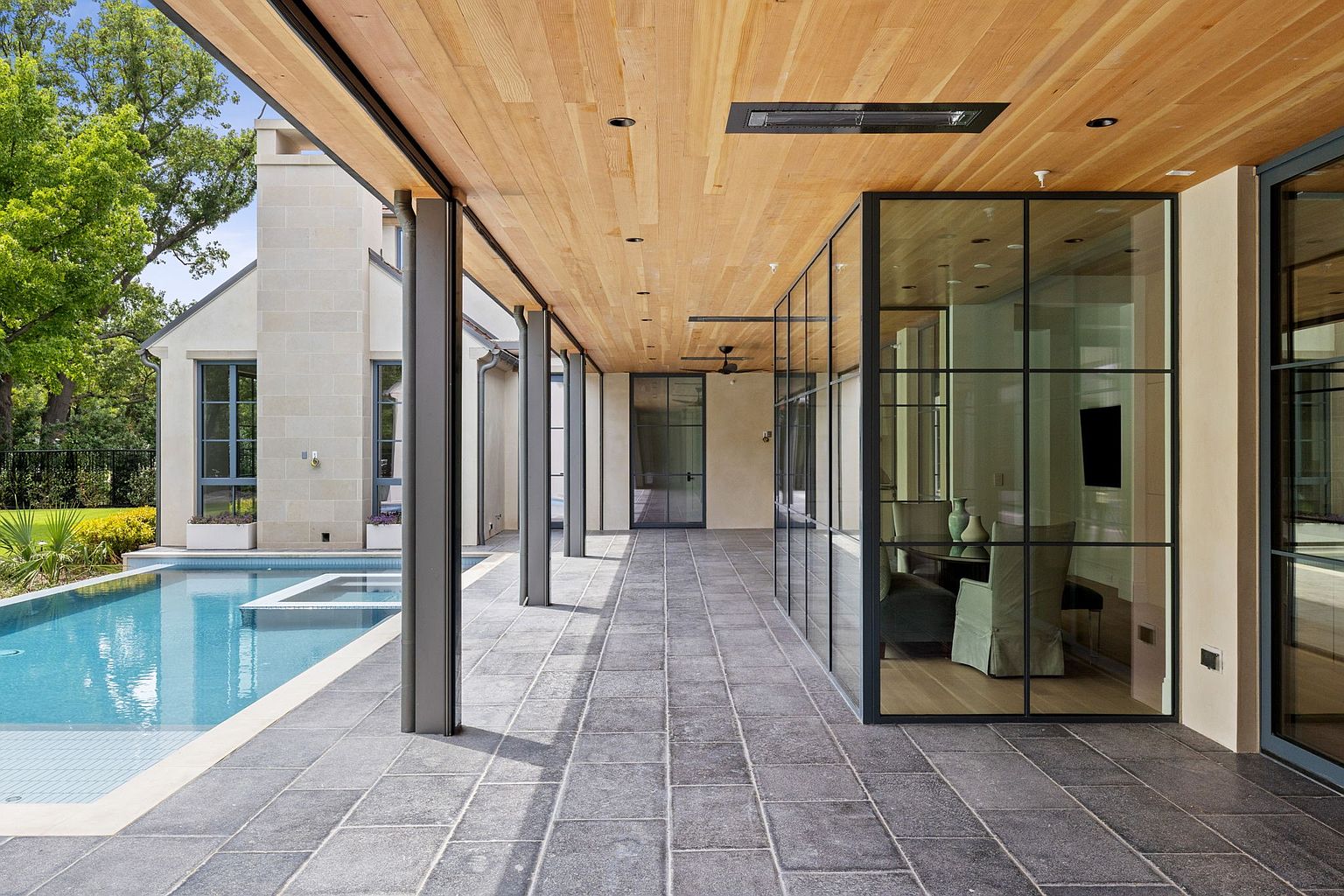 This image showcases a luxurious outdoor patio area featuring a sleek, modern design. The patio is covered with a wooden ceiling and supported by gray pillars, leading to a pool area. Large glass windows enclose an interior space, creating a seamless transition between indoor and outdoor living, while gray stone tiles cover the patio floor.