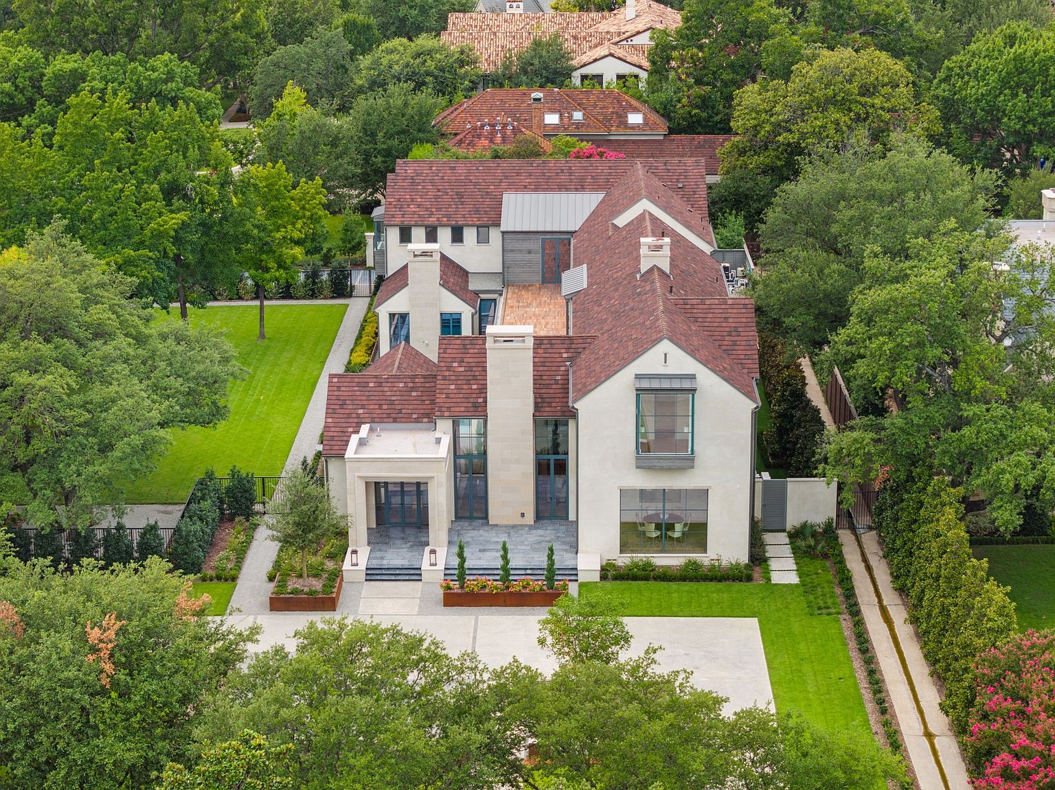 This aerial shot showcases a luxurious home with a red-tiled roof, set amidst lush greenery. The property features a well-manicured lawn, a paved driveway, and meticulously designed landscaping, creating an impression of refined elegance and privacy. The house's architectural details are highlighted from this vantage point, emphasizing its grandeur and curb appeal.