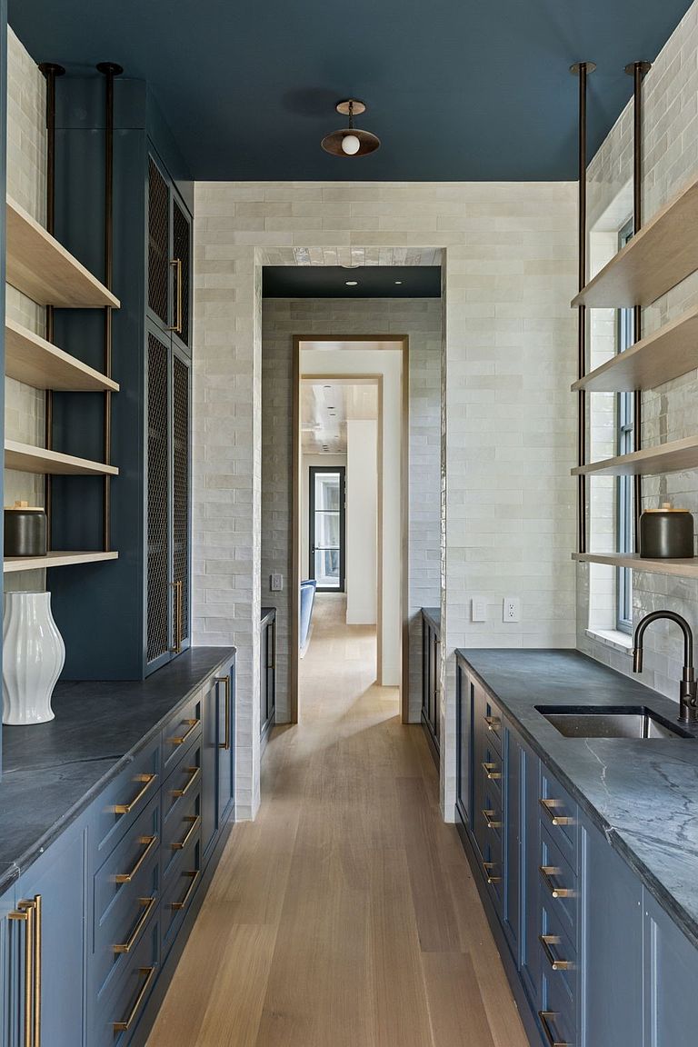This is an interior shot of a hallway featuring custom cabinetry and shelving on both sides. The cabinets are a muted blue with gold hardware, and the shelves are light wood. The walls are tiled in a light gray brick pattern, and the ceiling is painted a deep blue, creating a sophisticated and modern aesthetic.