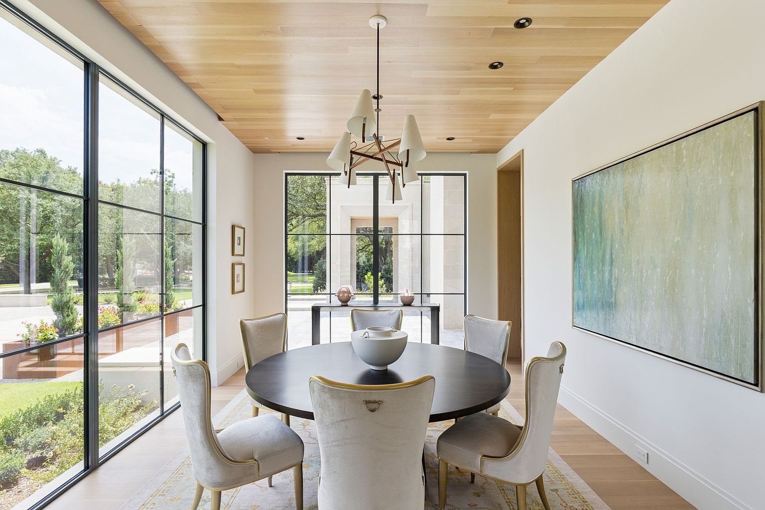 This is an interior shot of a dining room featuring a round, dark wood table surrounded by six upholstered chairs with gold accents. A modern chandelier hangs above the table, and a large window offers a view of the exterior landscape. An abstract painting adorns the wall, and the room is bathed in natural light, creating an elegant and inviting atmosphere.