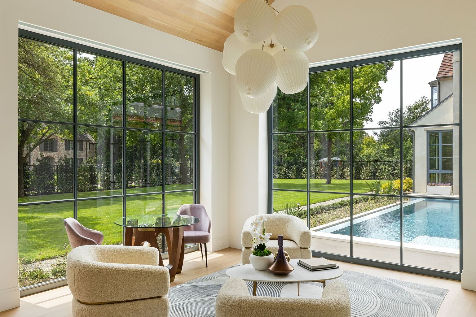 This is a bright and airy living room featuring large windows with black frames that offer stunning views of the lush green yard and pool. The room is furnished with comfortable, modern seating, including plush armchairs and a round coffee table, all arranged on a stylish area rug. A unique chandelier adds a touch of elegance to the space, creating a welcoming and sophisticated atmosphere.