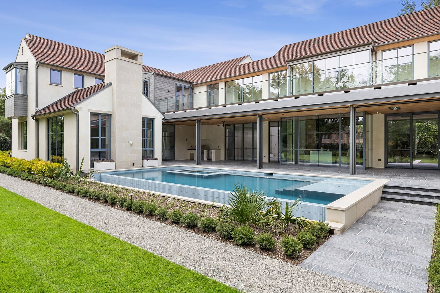 This image showcases the rear exterior of a luxurious home, emphasizing a modern pool and spa area. The pool features a unique design with a raised spa section, surrounded by manicured landscaping and a gravel pathway. The house itself boasts large windows and a covered patio area, creating a seamless indoor-outdoor living experience.