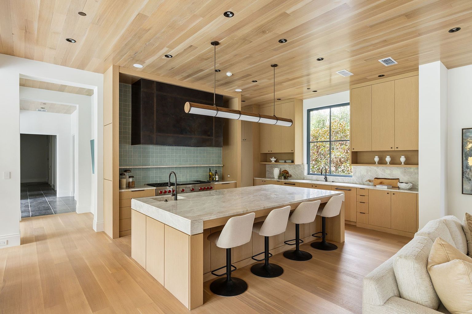 This is a bright and modern kitchen featuring light wood cabinetry, a large island with seating, and a dark metal range hood. The kitchen has a clean and minimalist design with a focus on natural materials and light. The perspective is from a wide angle, capturing the entire kitchen space and its connection to the adjacent living area.