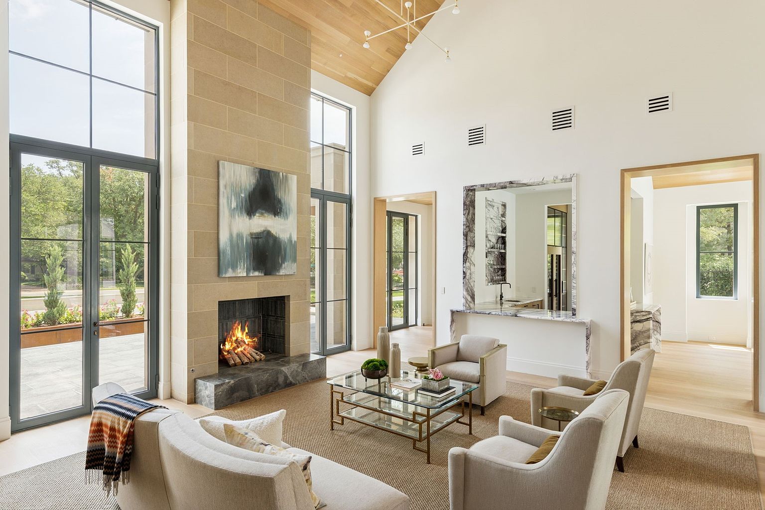 This is a bright and airy living room featuring a stone fireplace with a large abstract painting above it. The room is furnished with neutral-toned sofas and armchairs, arranged around a glass-topped coffee table. Large windows provide ample natural light and views of the outdoors, creating a sophisticated and inviting atmosphere.