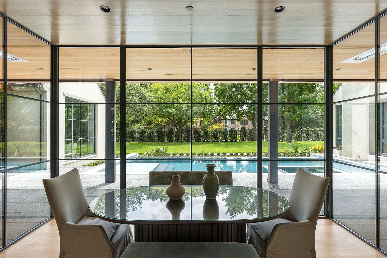This interior shot showcases a modern dining room with a glass-topped round table and upholstered chairs, set against a backdrop of floor-to-ceiling windows offering a view of a pool and landscaped yard. The room features a sleek, contemporary design with clean lines and natural light, creating an inviting and luxurious atmosphere. The perspective is from inside the room, looking out towards the pool area.
