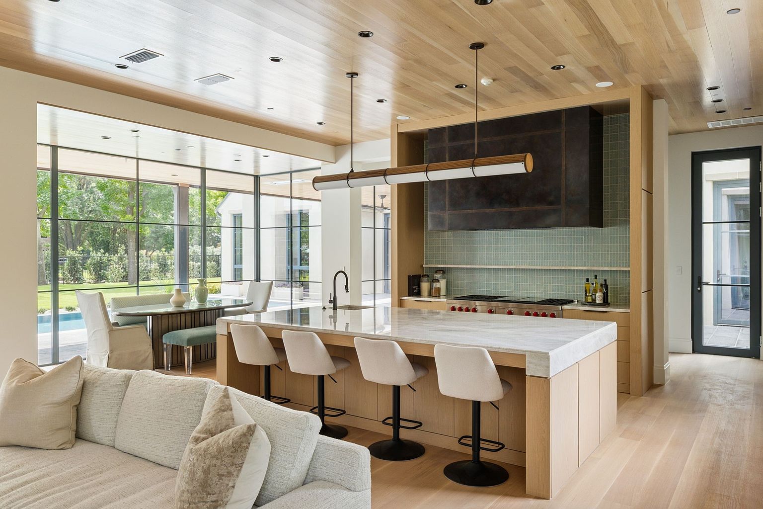 This is a bright and modern kitchen featuring a large island with bar seating, light wood cabinetry, and a striking dark metal range hood. The space is open to a dining area with large windows offering views of the outdoors, and the light wood flooring and ceiling create a warm and inviting atmosphere. A comfortable sofa is visible in the foreground, suggesting an open-concept living space.