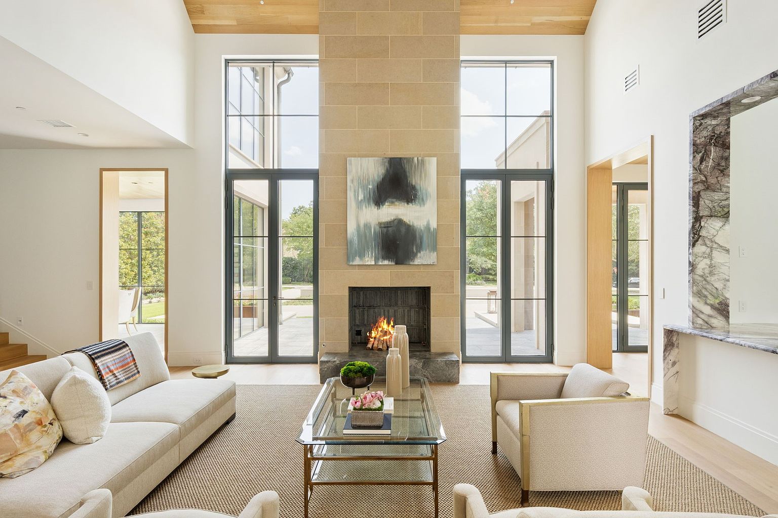 This is a spacious living room with a high ceiling and a large stone fireplace as the focal point. The room is well-lit with natural light coming through large windows on either side of the fireplace. The furniture includes a light-colored sofa, armchairs, and a glass coffee table, creating a comfortable and inviting atmosphere.