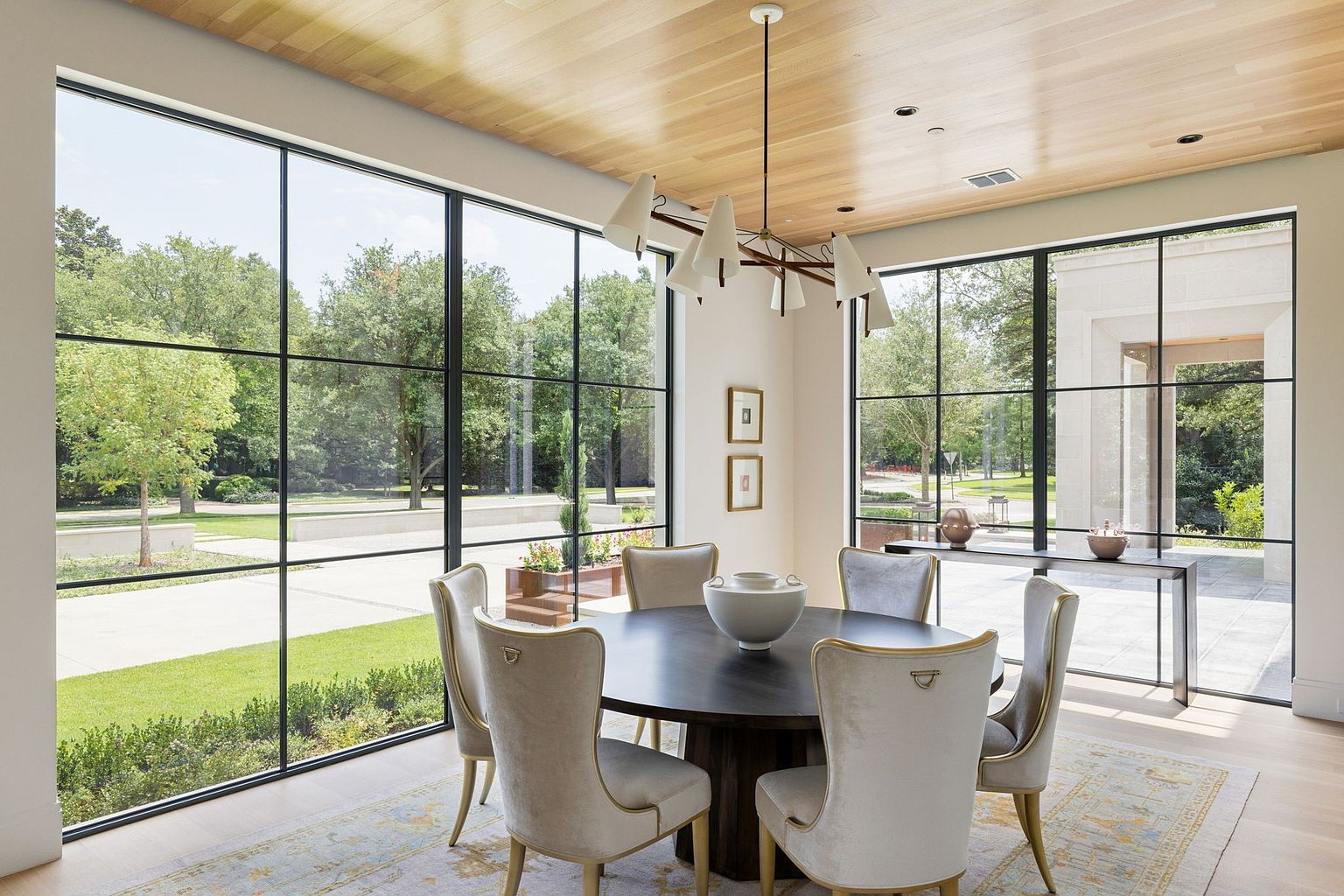 This is an interior shot of a dining room featuring a round dark wood table surrounded by six upholstered chairs with gold trim. A modern chandelier hangs above the table, and large windows offer a view of the landscaped yard. The room has a light and airy feel with a wood plank ceiling and a neutral color palette.