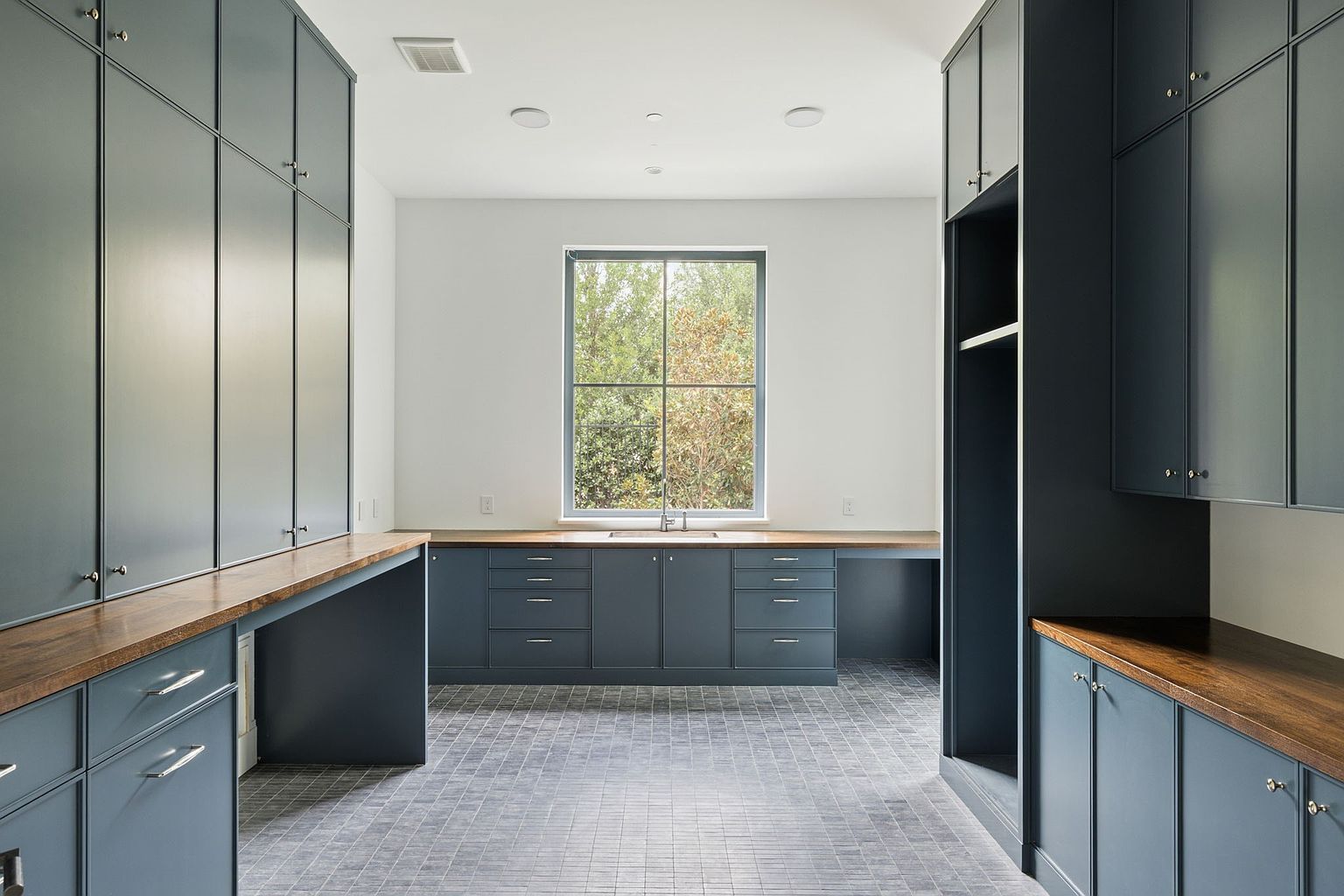 This is a well-organized laundry room featuring custom blue-gray cabinetry with wooden countertops, providing ample storage and workspace. A large window offers natural light, and the gray tiled floor adds a touch of modern elegance. The room appears clean, functional, and stylish, making it an attractive feature for potential buyers.