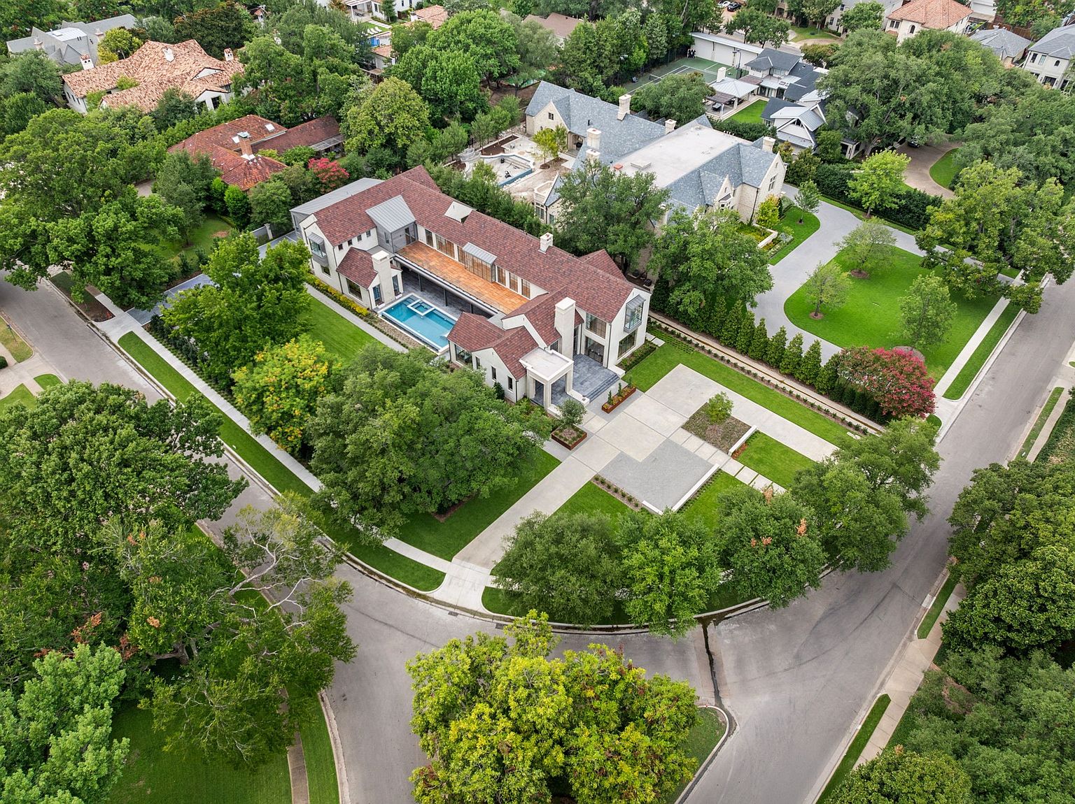 This aerial view showcases a luxurious estate with a modern architectural design, featuring a red-tiled roof, a private pool, and meticulously landscaped grounds. The property includes a spacious driveway, mature trees, and manicured lawns, creating an impression of exclusivity and privacy. The surrounding neighborhood is lush and residential, adding to the overall appeal.
