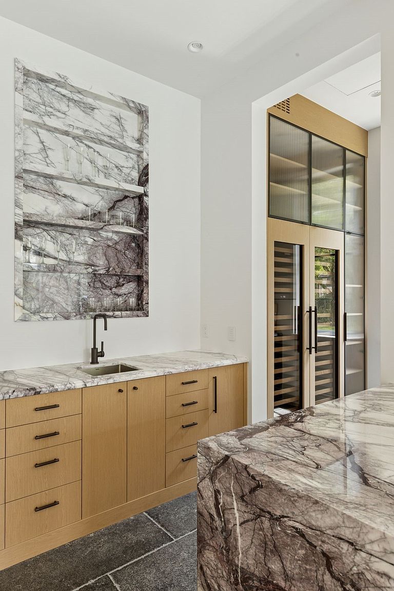 This interior shot showcases a modern kitchen area with light wood cabinetry, marble countertops, and a built-in wine refrigerator. A decorative marble backsplash with shelving adds a unique touch, while the dark hardware provides a sleek contrast. The overall design exudes sophistication and functionality.