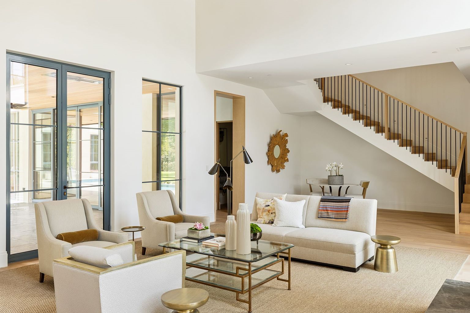 This is an interior shot of a living room featuring a neutral color palette with beige sofas and armchairs, a glass and gold coffee table, and a large area rug. The room has high ceilings and large windows that provide ample natural light. A staircase is visible in the background, adding architectural interest.