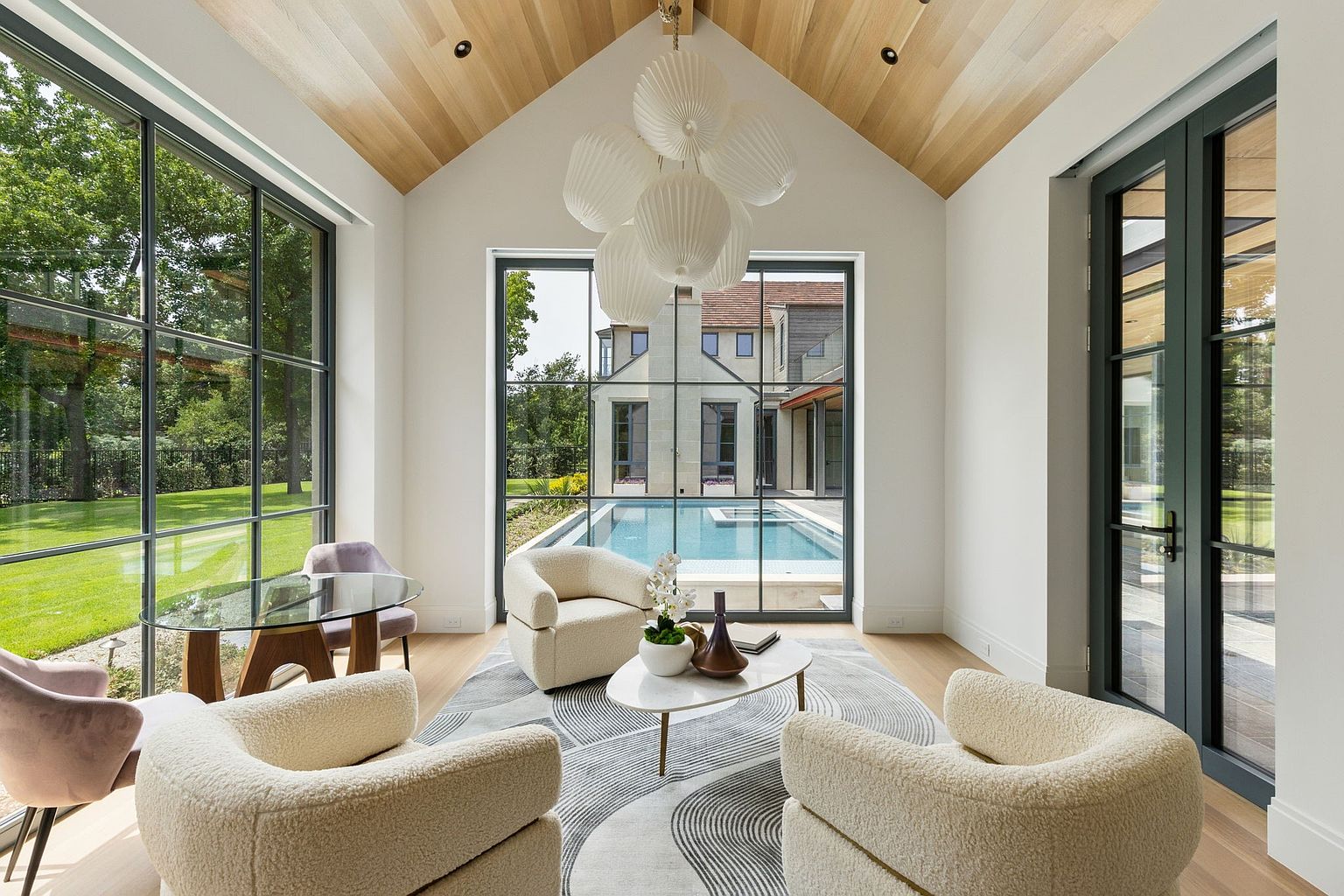 This is an interior shot of a modern living room featuring high ceilings with wooden panels and large windows offering a view of the pool and backyard. The room is furnished with plush, curved armchairs, a round glass-top table, and a patterned rug, creating a bright and inviting space. The color palette is neutral with pops of color from the outdoor greenery and the lavender chairs.