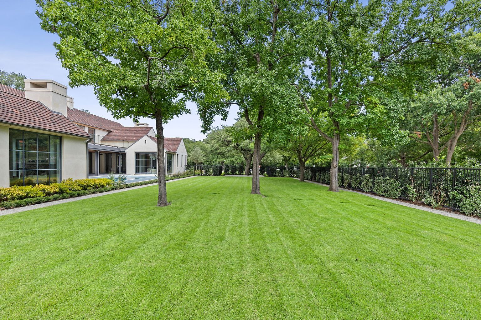 This image showcases a meticulously maintained backyard featuring a lush green lawn, mature trees providing shade, and a well-defined border with manicured hedges and a black metal fence. The property includes a modern house with large windows and a red tile roof, suggesting a luxurious and private outdoor space perfect for relaxation and entertainment. The perspective is a wide shot, emphasizing the expanse of the yard.