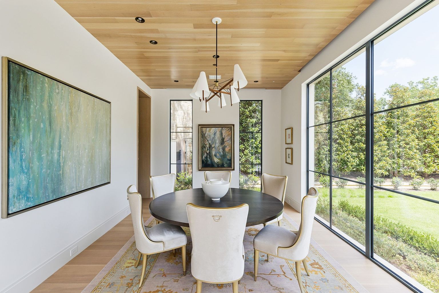 This is an interior shot of a dining room featuring a round dark wood table surrounded by six upholstered chairs with gold accents. A modern chandelier hangs above the table, and large windows offer a view of the lush greenery outside. Artwork adorns the walls, and a patterned rug anchors the space, creating an elegant and inviting atmosphere.