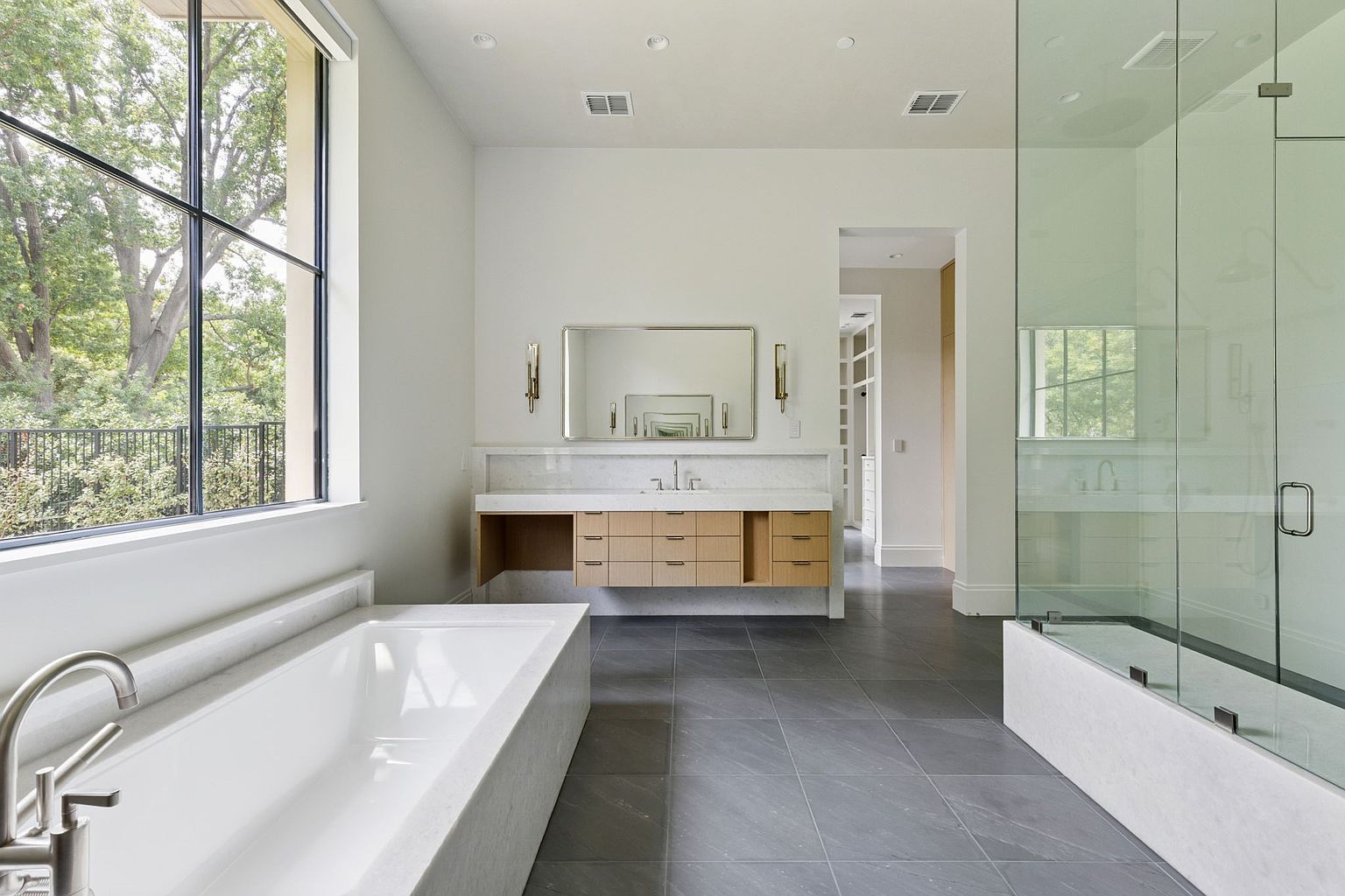 This is a luxurious primary bathroom featuring a large soaking tub next to a window, a modern vanity with a marble countertop and wooden drawers, and a glass-enclosed shower. The bathroom has gray tile flooring and neutral-colored walls, creating a serene and spa-like atmosphere. The perspective is a wide shot, showcasing the spaciousness and high-end finishes.