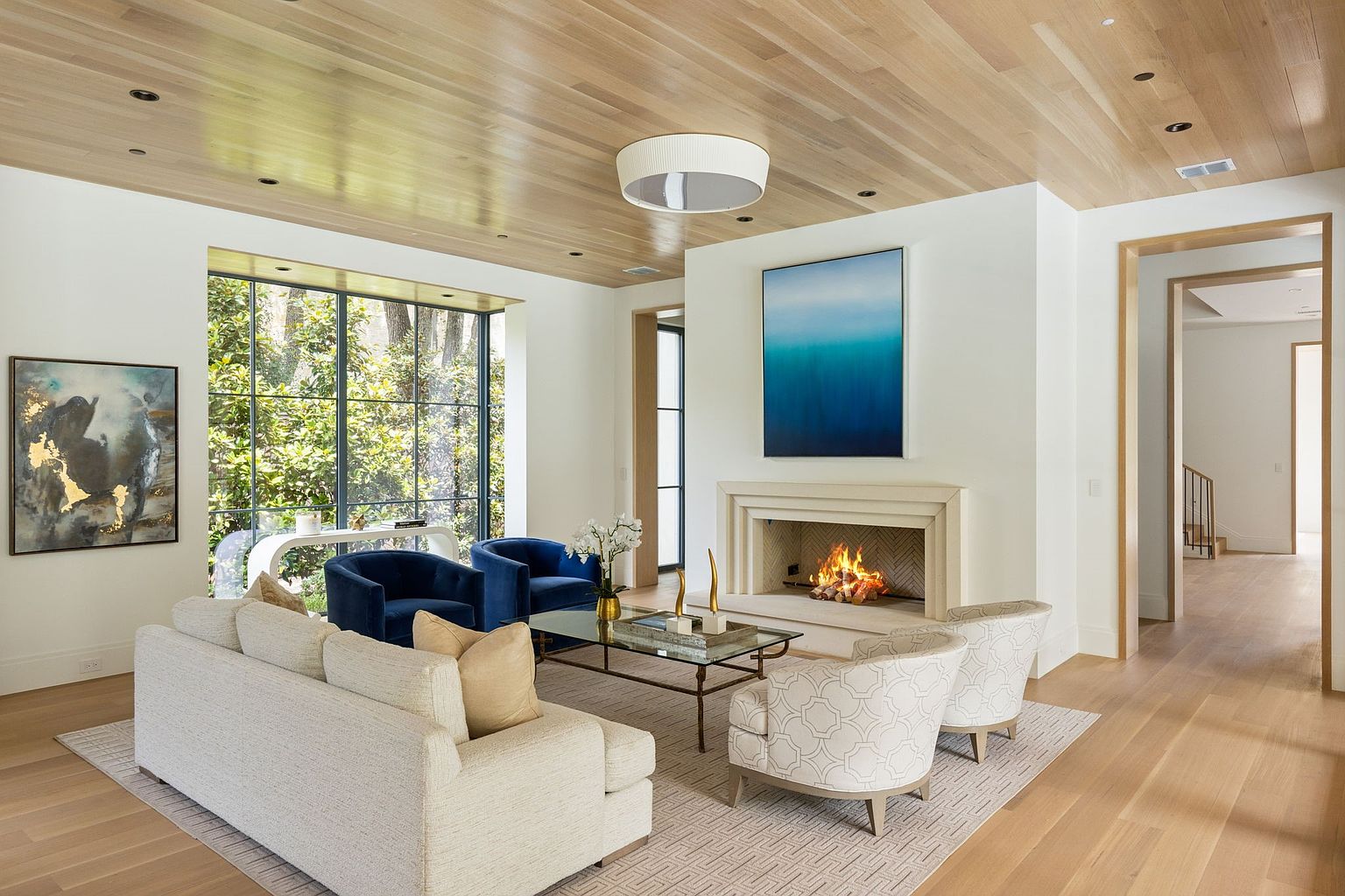 This is an interior shot of a living room featuring a fireplace, a light-colored sofa with throw pillows, and two blue armchairs. The room is well-lit with natural light coming through a large window, and the ceiling is made of wood. The overall style is modern and elegant, creating a warm and inviting atmosphere.