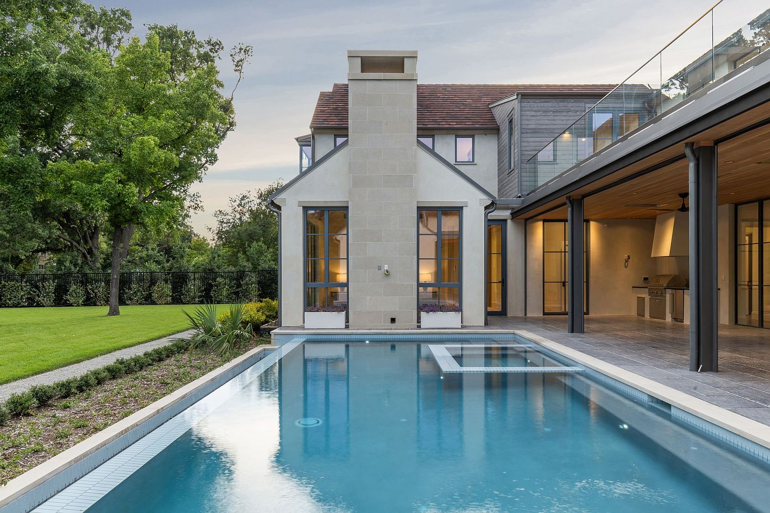 This image showcases the rear exterior of a luxurious home, emphasizing a stunning pool and spa area. The pool features a modern design with clean lines and a built-in spa. The home's architecture includes a stone chimney, large windows, and a covered patio with an outdoor kitchen, creating an inviting space for relaxation and entertainment. The perspective is from the pool level, looking towards the house, creating a sense of immersion and highlighting the seamless integration of indoor and outdoor living.