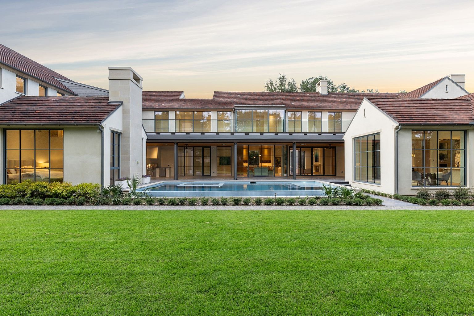 This is a rear view of a luxurious home featuring a large swimming pool, expansive lawn, and a modern architectural design. The house has large windows and doors that open to the pool area, creating a seamless indoor-outdoor living experience. The exterior is characterized by clean lines, a neutral color palette, and a well-manicured landscape, enhancing the property's appeal.