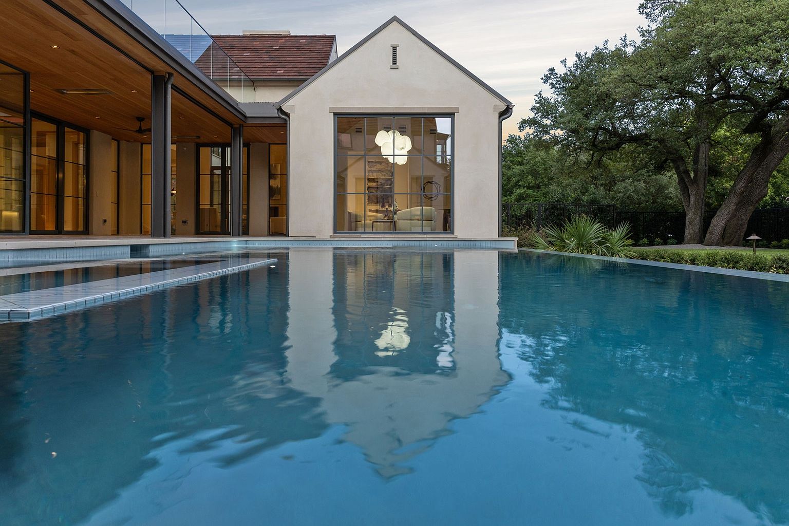 This image showcases a luxurious pool area of a modern home. The pool's calm, blue water reflects the architecture of the house, including a large window and the building's facade. The setting is serene, with mature trees visible in the background, suggesting a private and upscale residential environment.