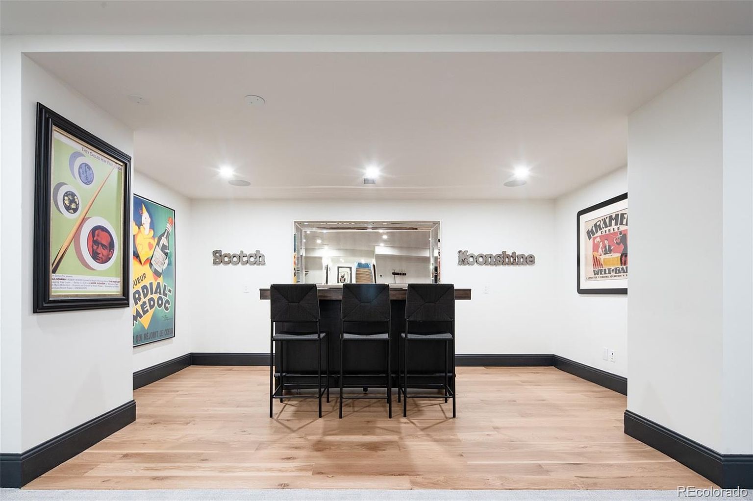 This is an interior shot of a basement bar area. The bar features a dark wood countertop with three black bar stools. Above the bar is a mirror, flanked by the words "Scotch" and "Moonshine." Two framed posters hang on the walls, and the flooring is light wood. The room is well-lit with recessed lighting, creating a clean and inviting space.